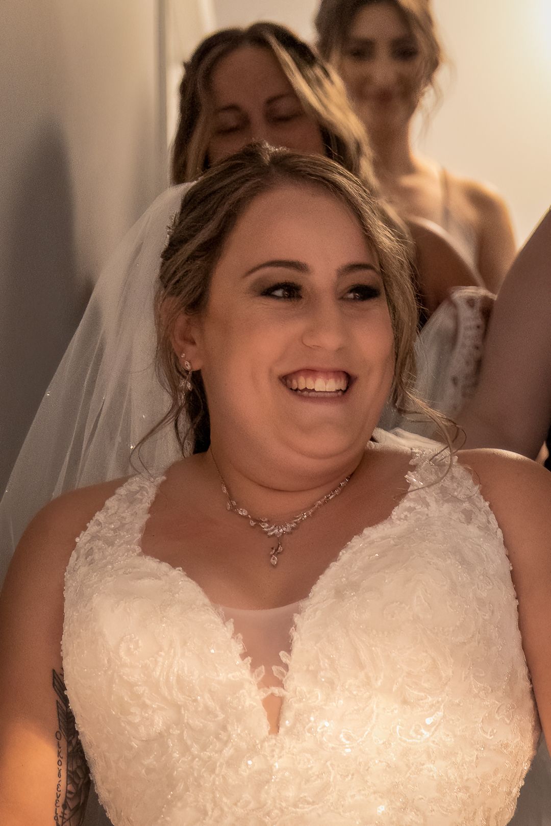 Smiling bride in white lace wedding gown and veil with diamond necklace, bridesmaids visible in background.