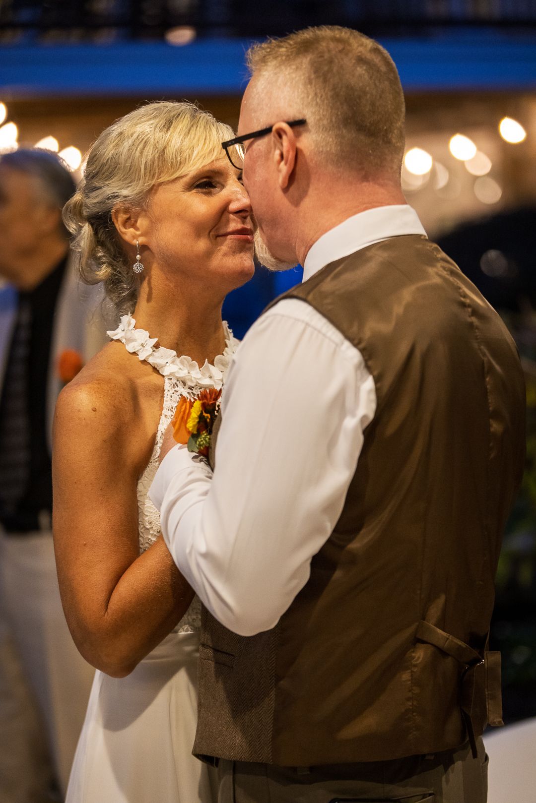 Couple sharing first dance at wedding reception, bride in white dress and groom in brown vest, string lights glowing behind them.