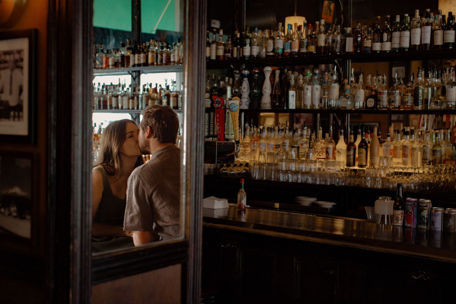 A man and woman share a kiss in a mirror in front of a bar filled with colorful liquor bottles.