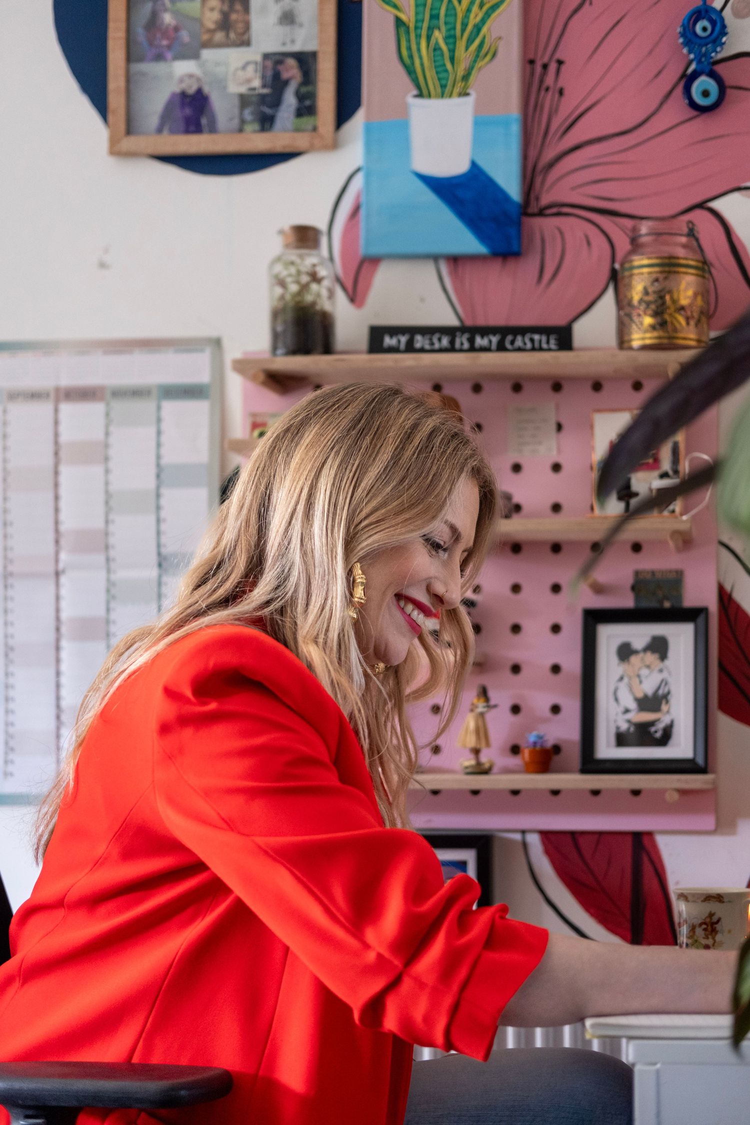Smiling blonde woman in red blazer working at a colorful creative desk with pegboard and artwork in a stylish home office.