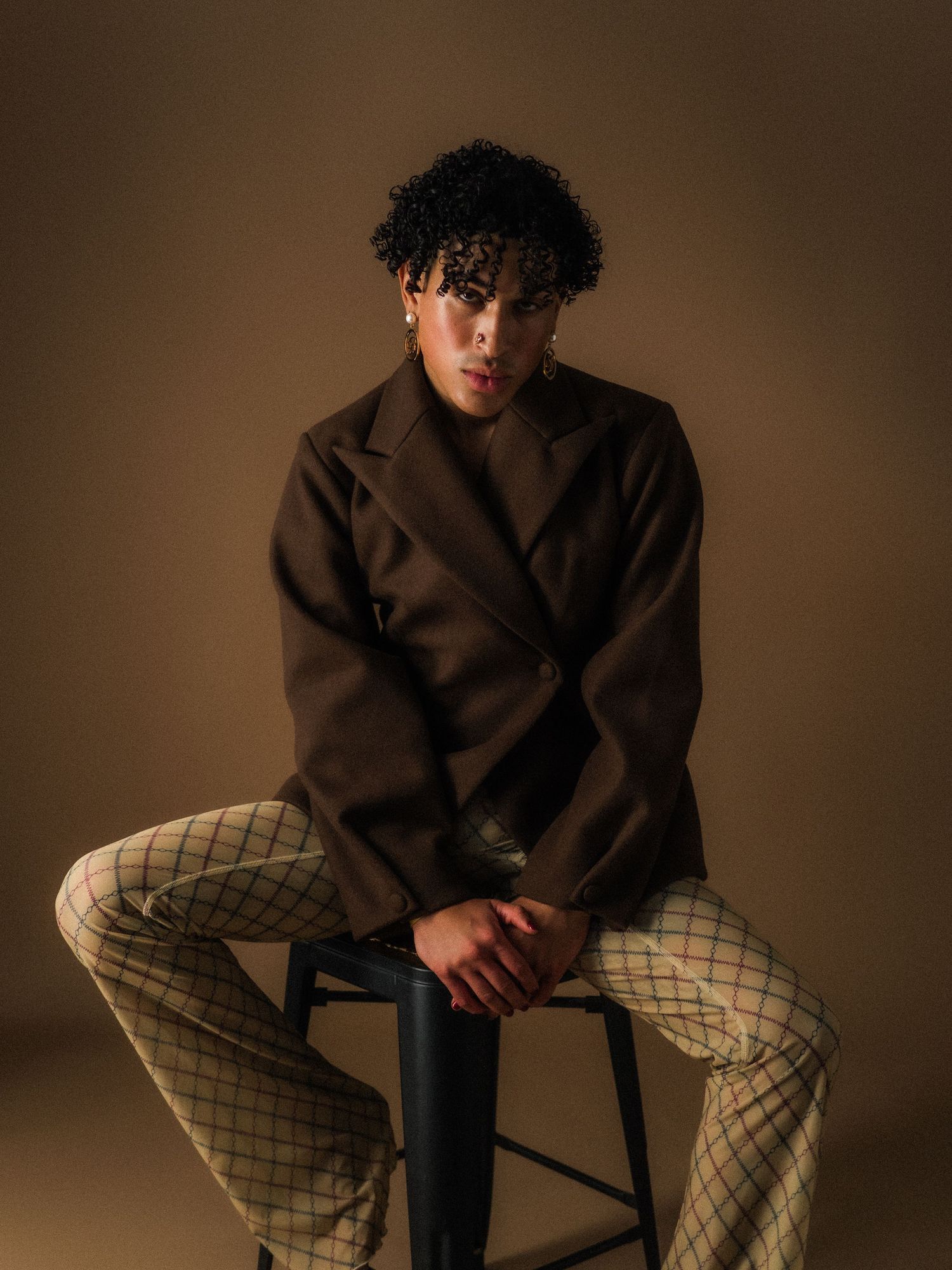 Young man with curly hair sits on stool wearing brown blazer and plaid trousers against warm beige backdrop.