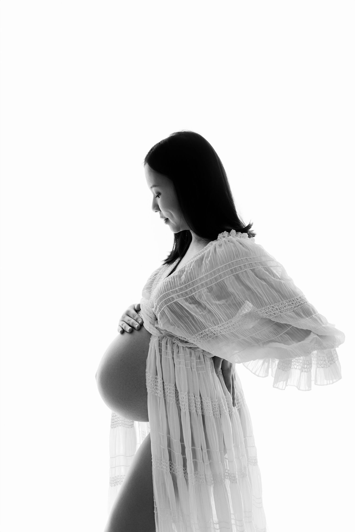 Black and white maternity photo of a pregnant woman in a lace robe cradling her baby bump.