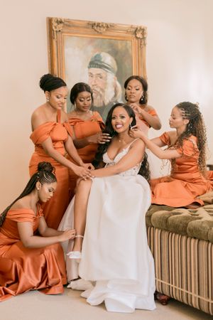 Bride in white gown surrounded by four bridesmaids in orange satin dresses helping her get ready indoors.