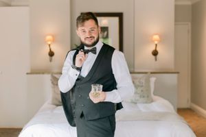 Dapper groom in black vest and bow tie holds whiskey glass in elegant hotel room.