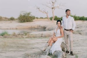 Elegant couple poses on rocky terrain with baobab trees and dry landscape in background.
