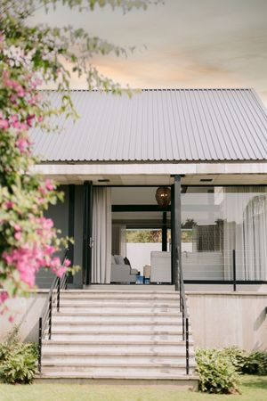 Modern venue exterior with concrete stairs, metal roof, glass walls, and pink bougainvillea framing the entrance at dusk.