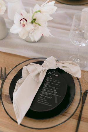 Elegant wedding place setting with black plate, cream bow napkin, menu card, and white orchids on wooden table.