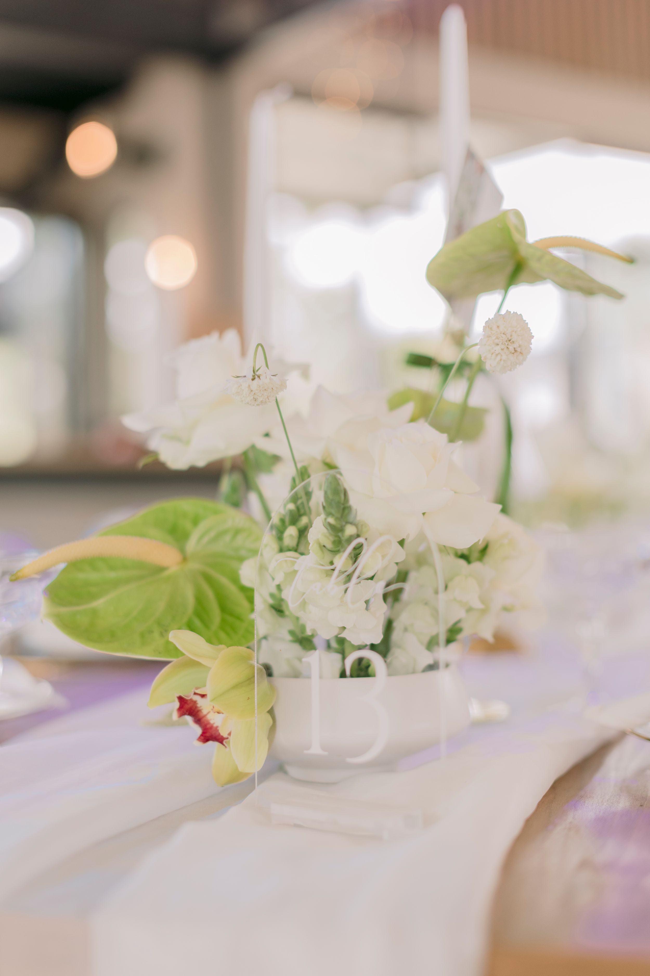 Elegant wedding table centerpiece with white flowers and green tropical leaves in a glass vase on a white tablecloth.