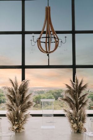 Elegant wedding ceremony setup with pampas grass arrangements, acrylic podium, wood chandelier, and large windows at sunset.