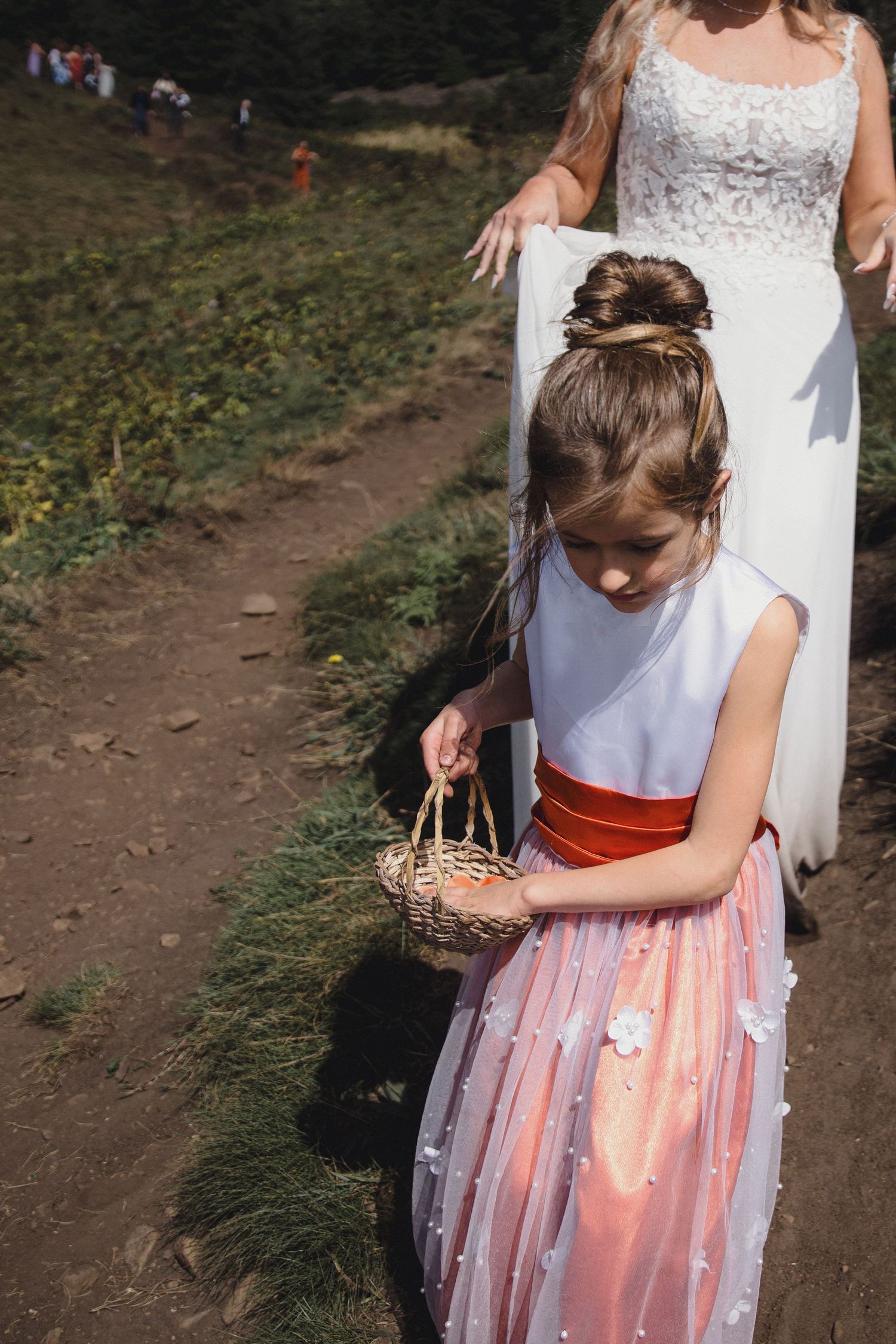 Young flower girl in white and pink dress carries basket while walking with bride on outdoor path.