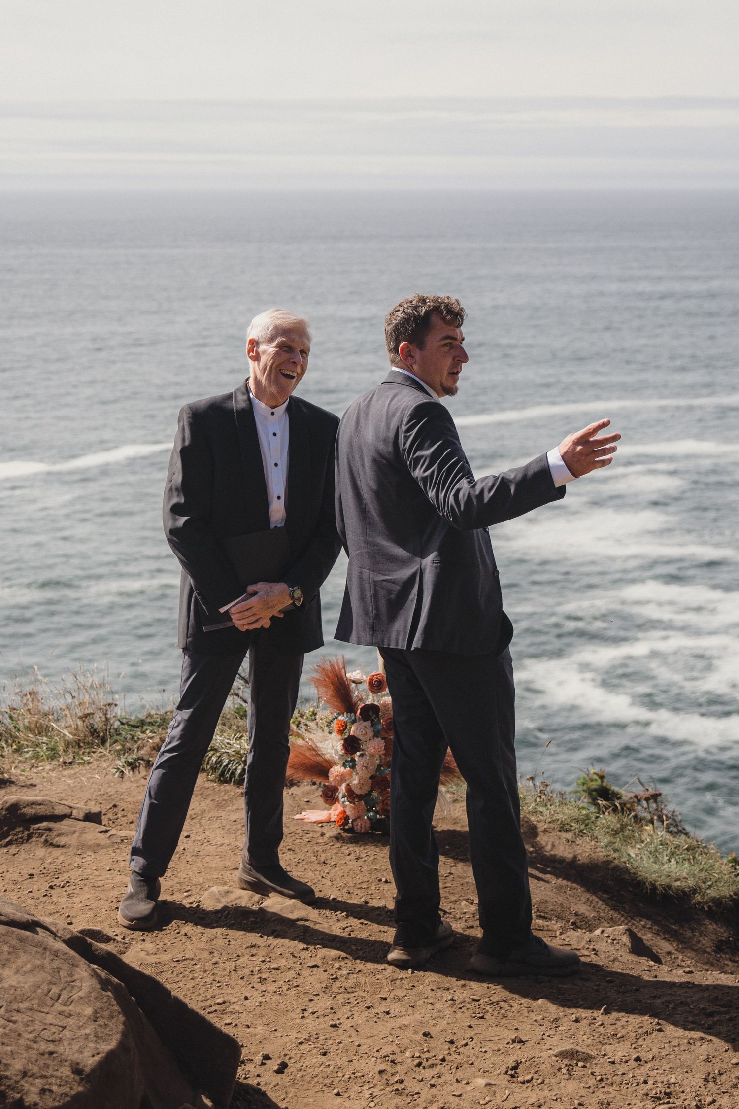 Two men in suits stand on a coastal cliff overlooking the ocean at an outdoor wedding ceremony.