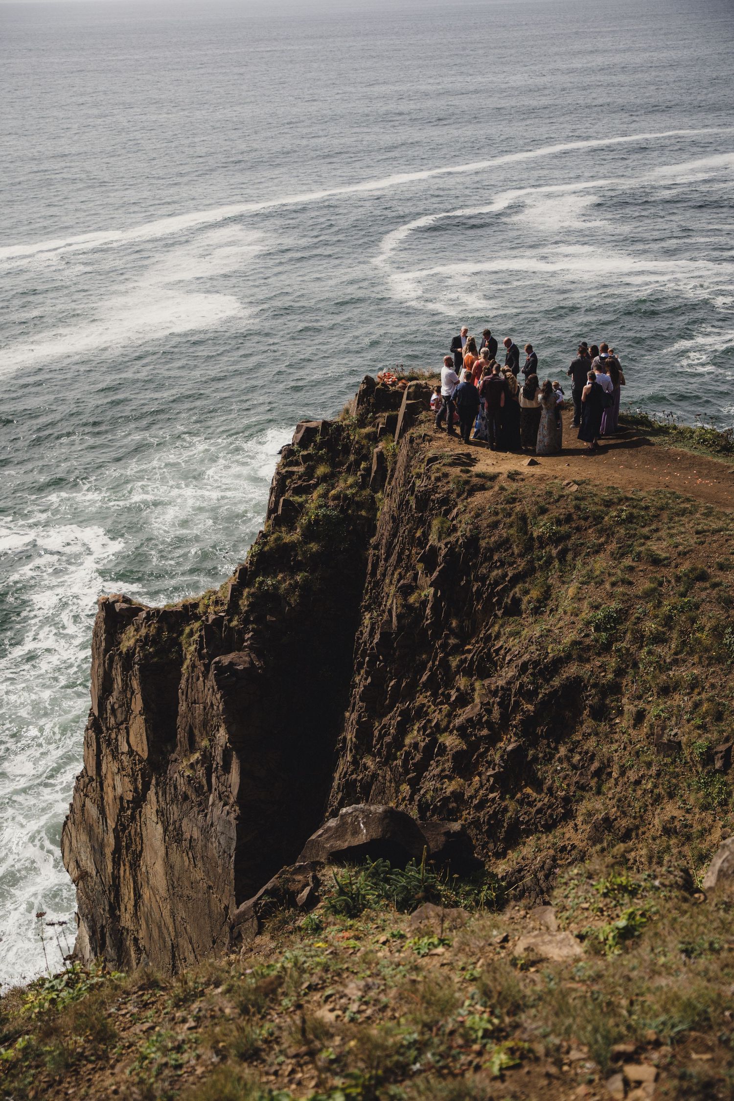Group of people gathered on a dramatic coastal cliff edge overlooking crashing ocean waves below.