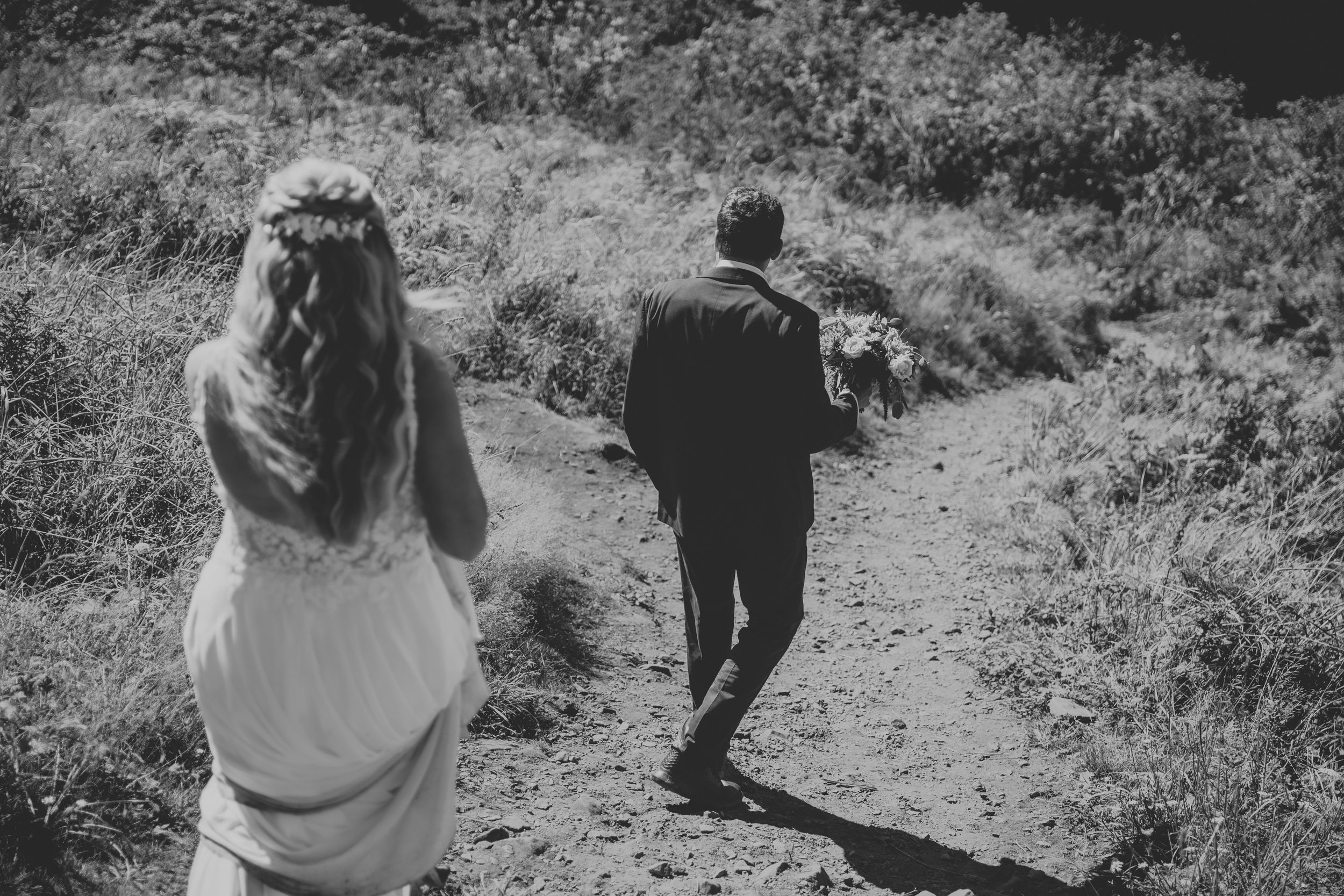Black and white photo of a bride and groom walking hand in hand along a rustic dirt path surrounded by lush greenery.