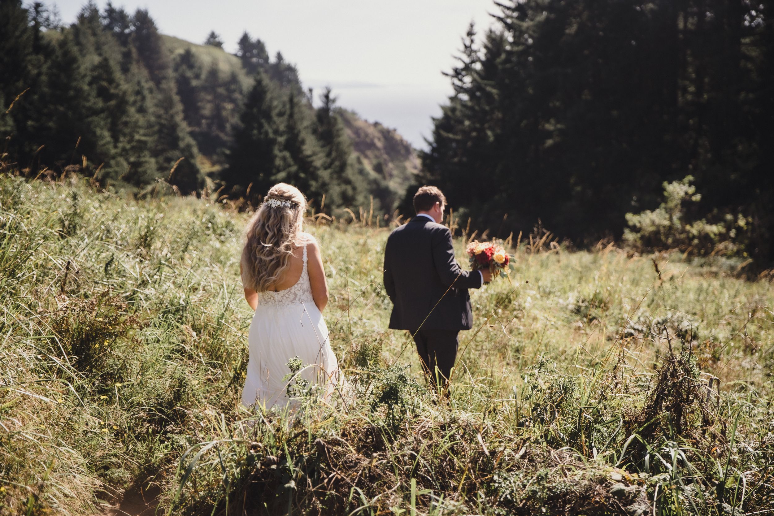 Bride and groom walking through a lush mountain meadow surrounded by tall evergreen trees on their wedding day.