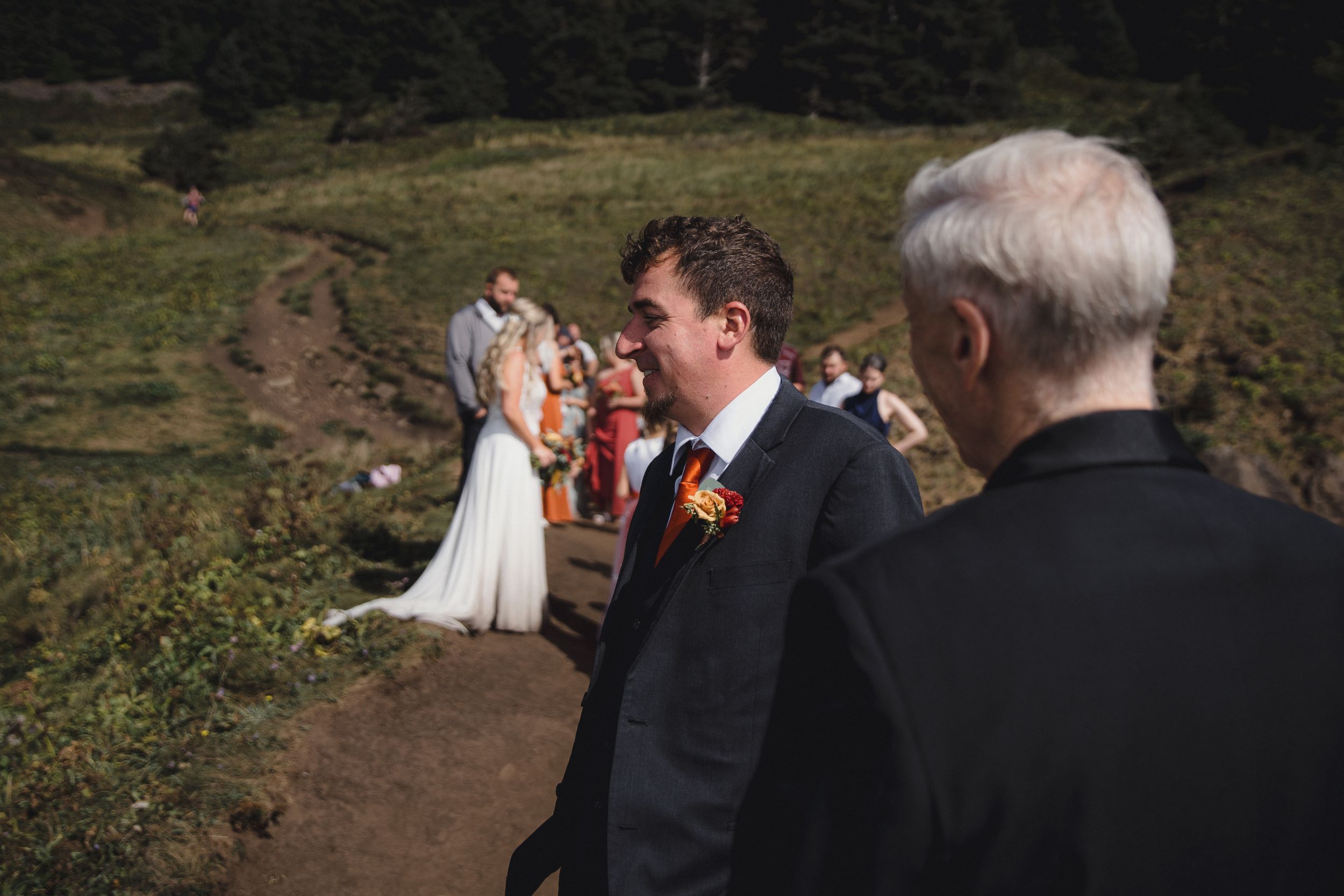 Groom in dark suit with orange boutonniere smiles during outdoor wedding ceremony on scenic hillside.