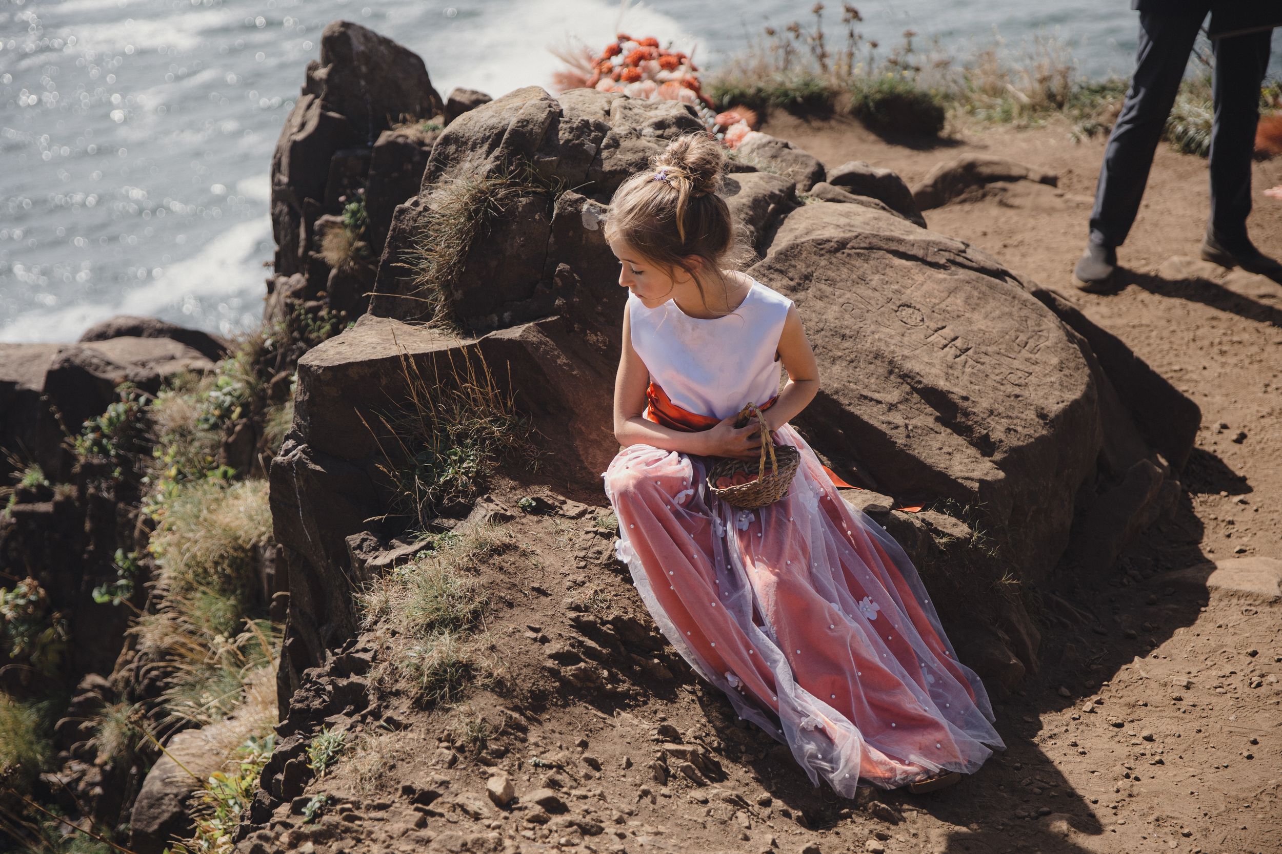 Woman in white top and pink floral skirt sitting on rocky coastal cliff overlooking the ocean.