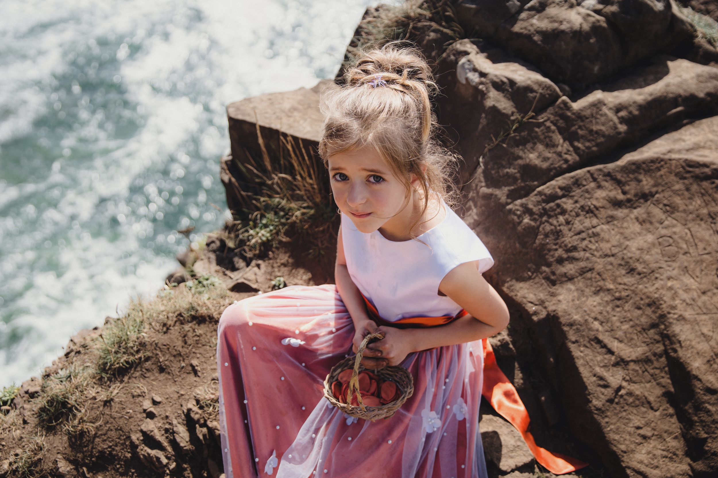 Young girl in pink skirt and white top sitting on rocky coastal cliffs with ocean waves in background.