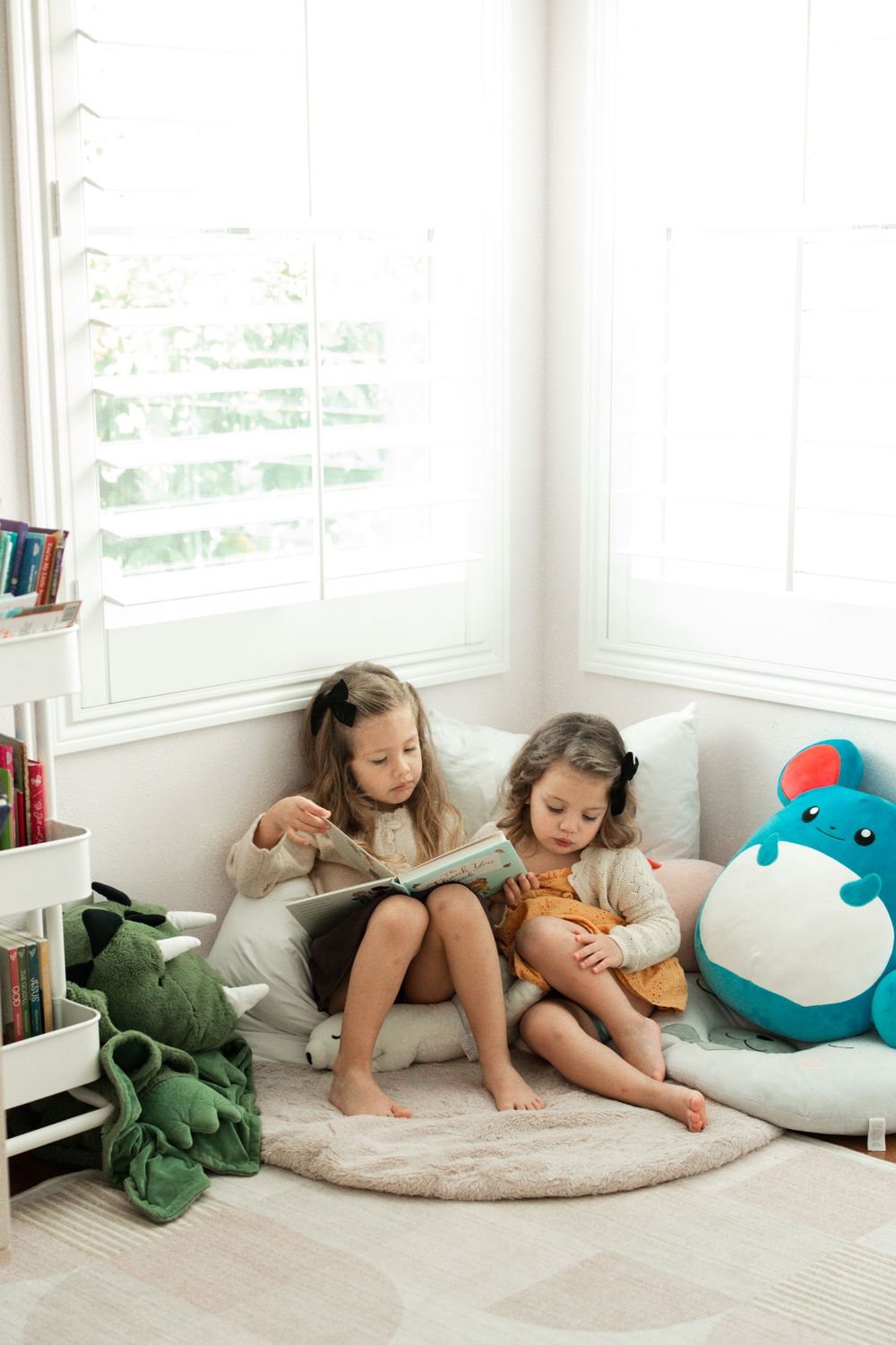 Two young girls sitting on a round rug reading a book together, surrounded by colorful stuffed animals.