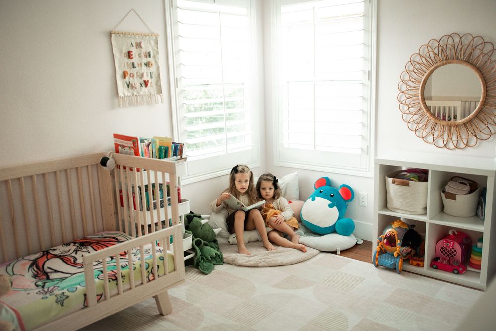 Two young girls sit on a round rug in a bright nursery with a crib, toy shelf, and rattan mirror.