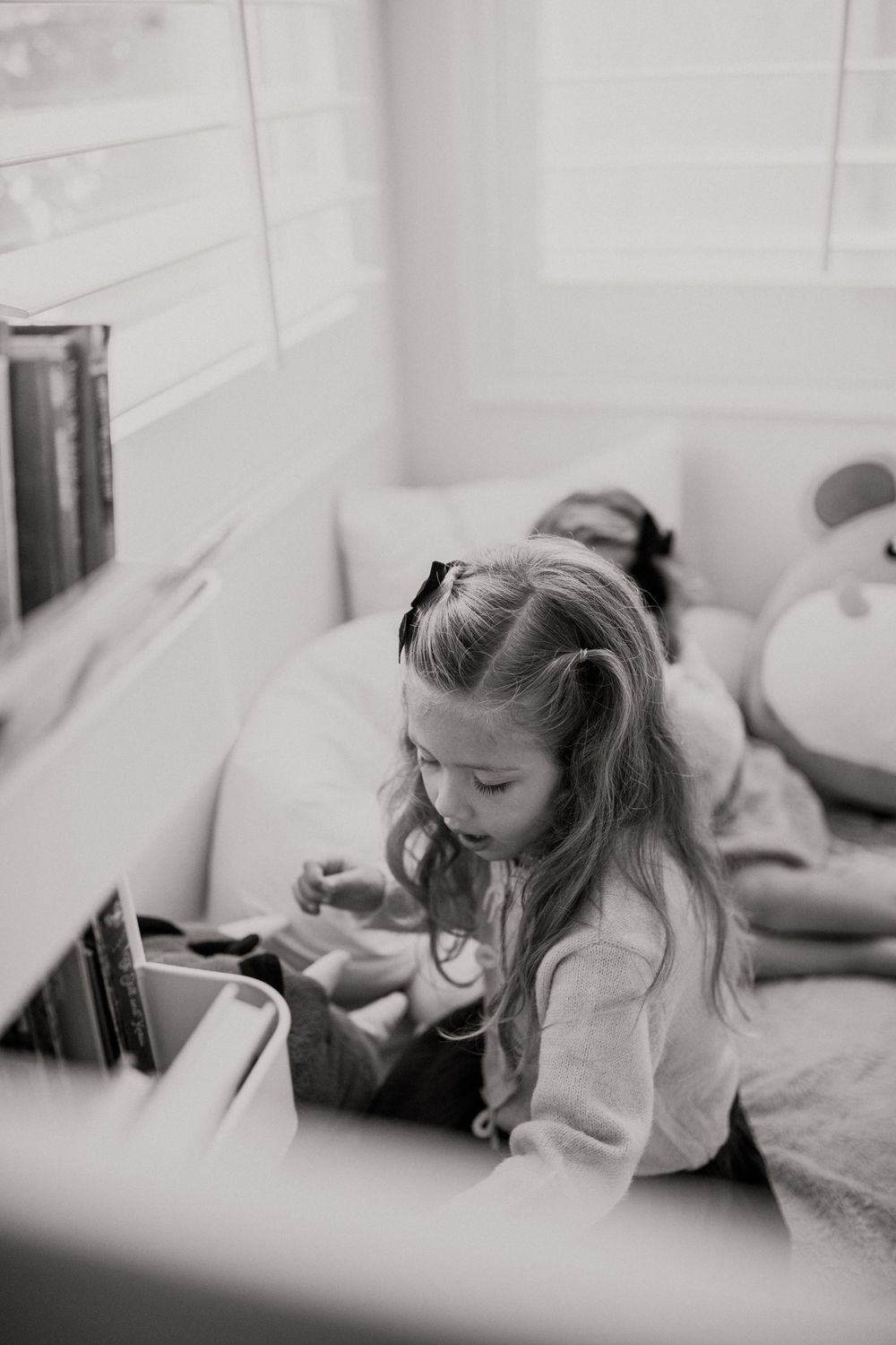 A young girl with long hair reads quietly on a cozy window seat beside a bookshelf, in black and white.