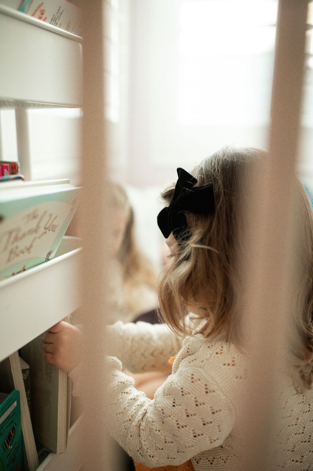 A young girl with a black bow hair clip browses books on a white shelf in soft natural light.