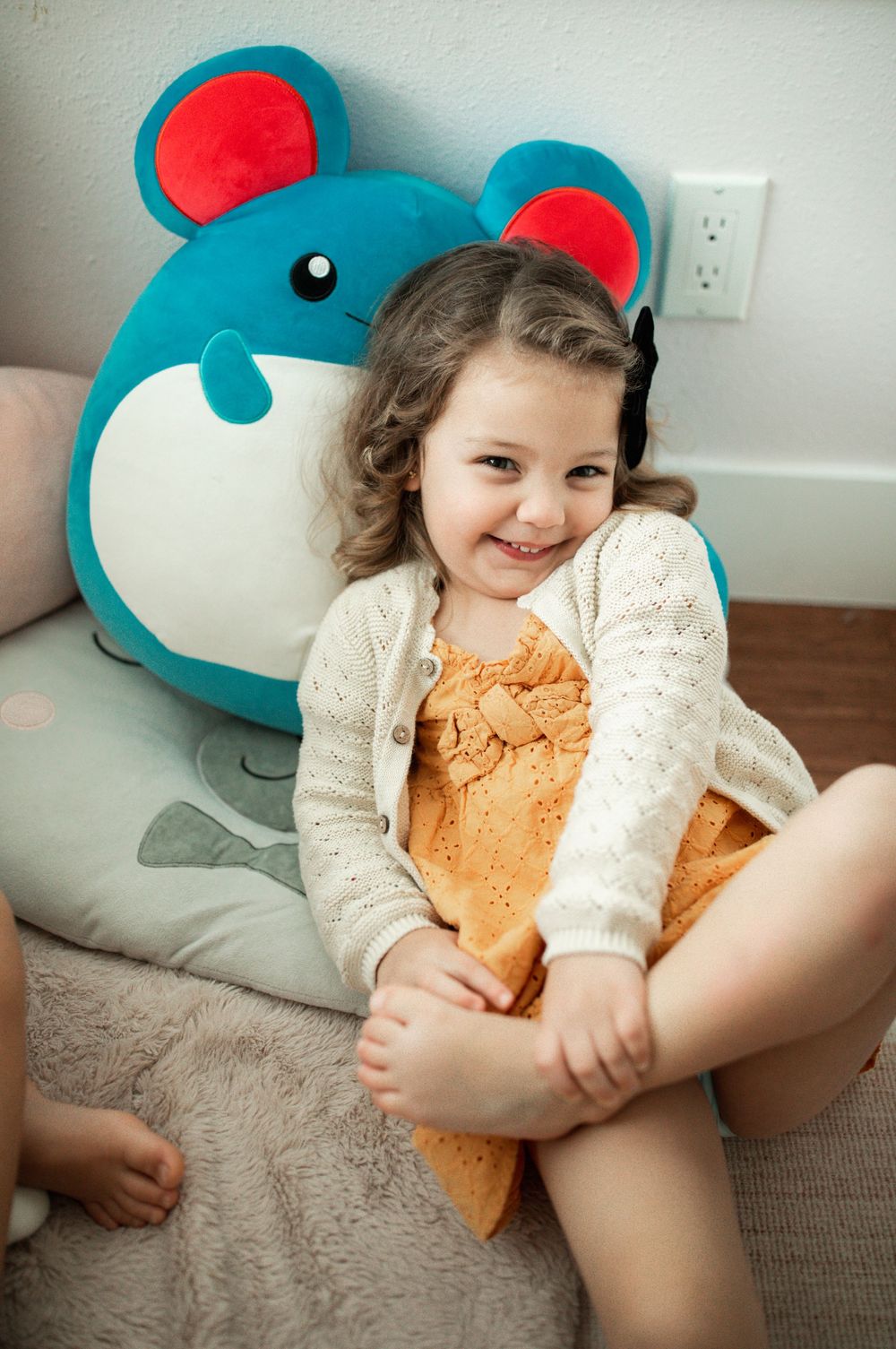 A smiling toddler girl in an orange dress and white cardigan sits against a colorful mouse plush toy.