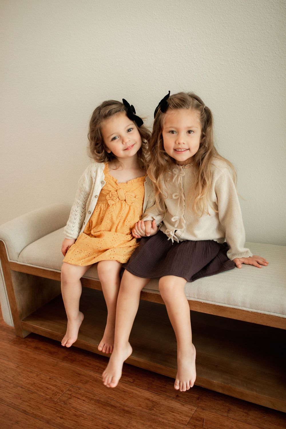Two young girls in matching black bows sit together on a bench, one in orange dress, one in beige top and dark skirt.
