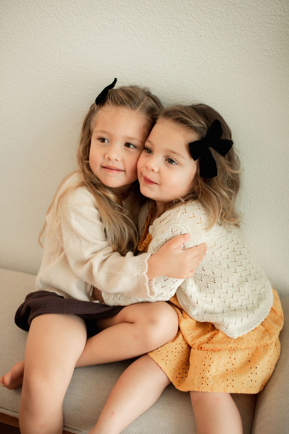 Two young sisters in knit sweaters and black hair bows share a sweet hug while sitting together.