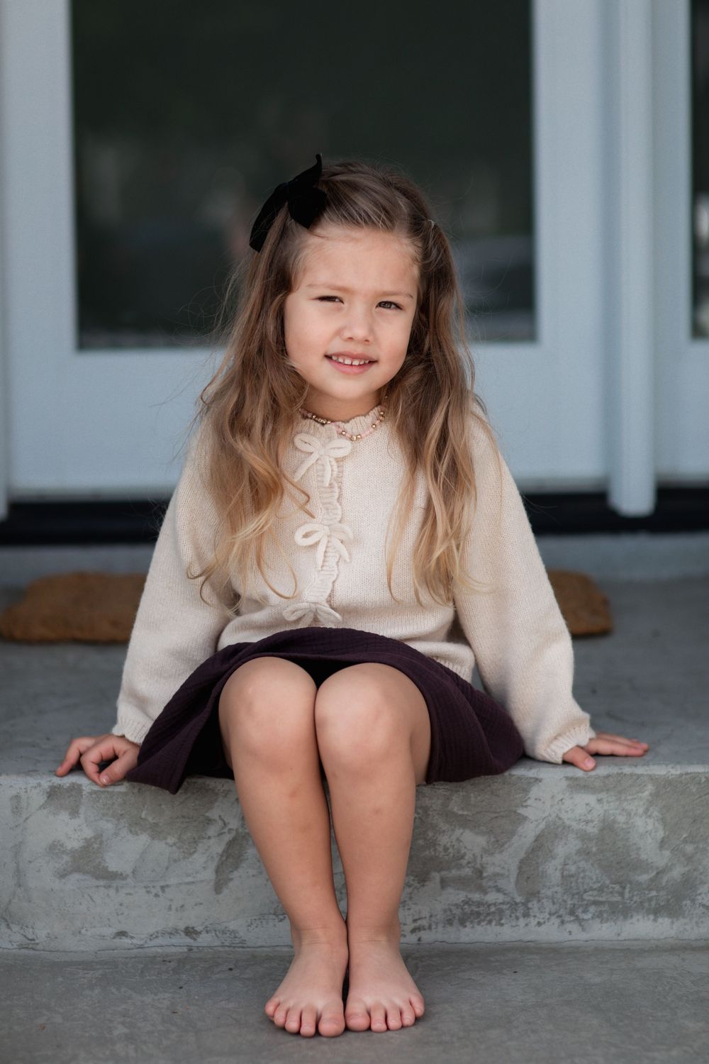 A smiling young girl with long hair and a black bow sits barefoot on steps in a cream sweater and dark skirt.