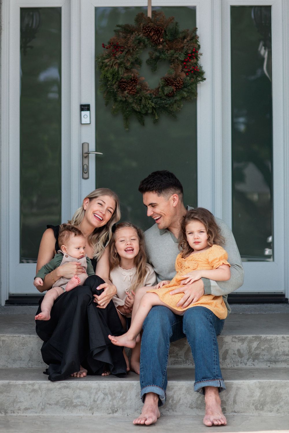 Happy family of five sitting on front porch steps with Christmas wreath on door behind them.