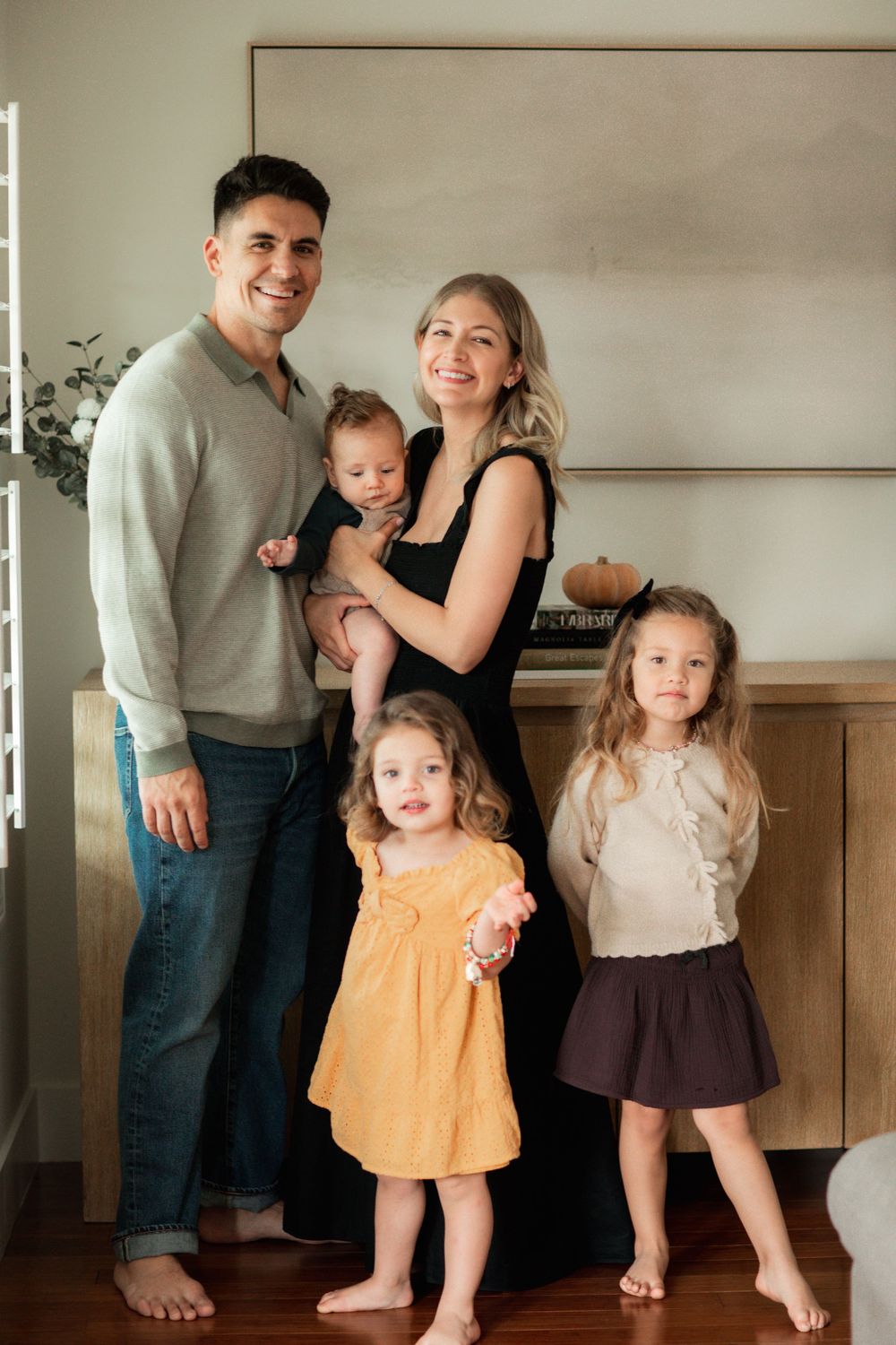 Family of five posing indoors, parents holding baby with two young girls standing in front.