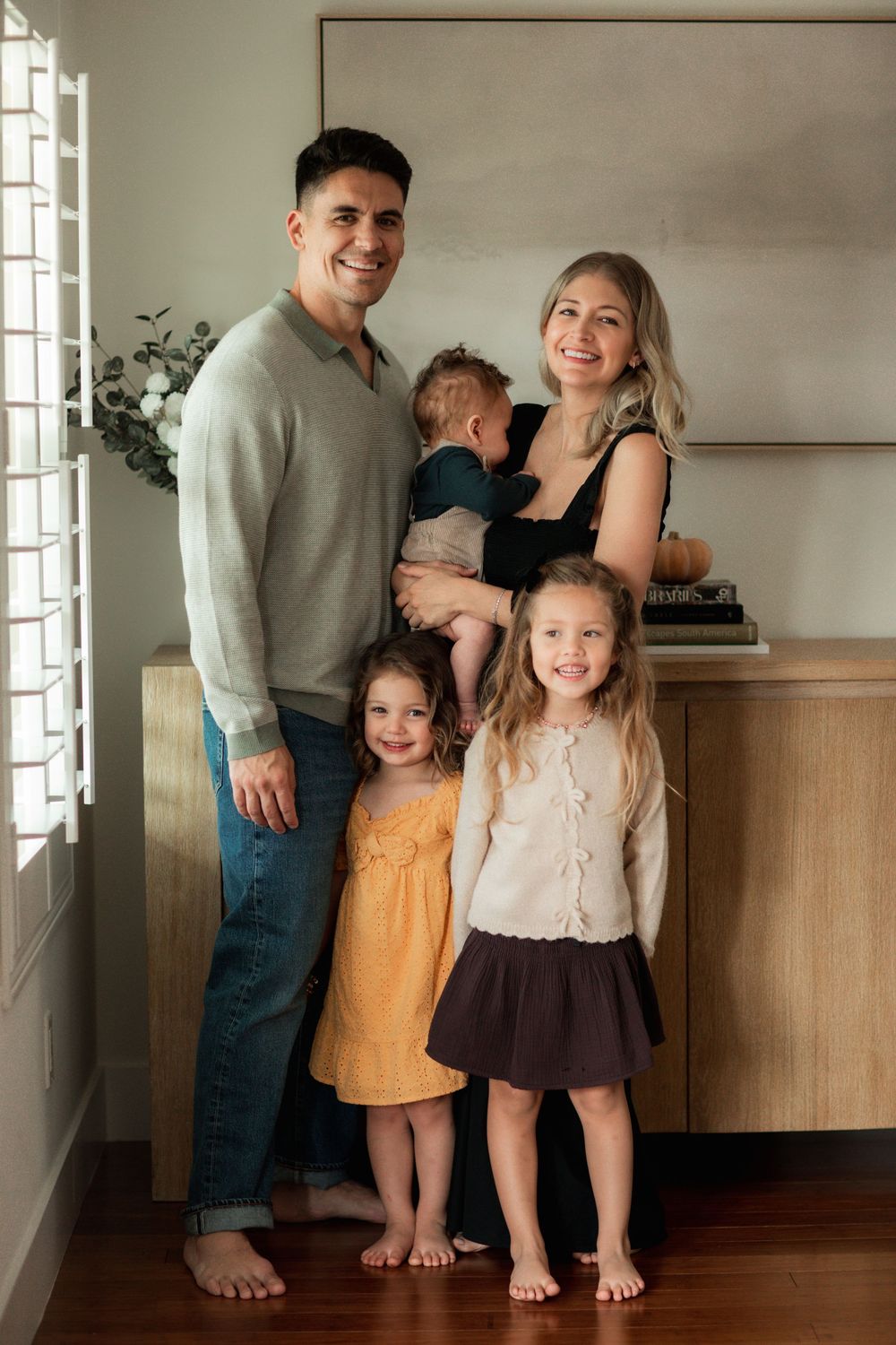 A smiling family of five poses indoors, parents with three young children including a baby, dressed in casual autumn attire.