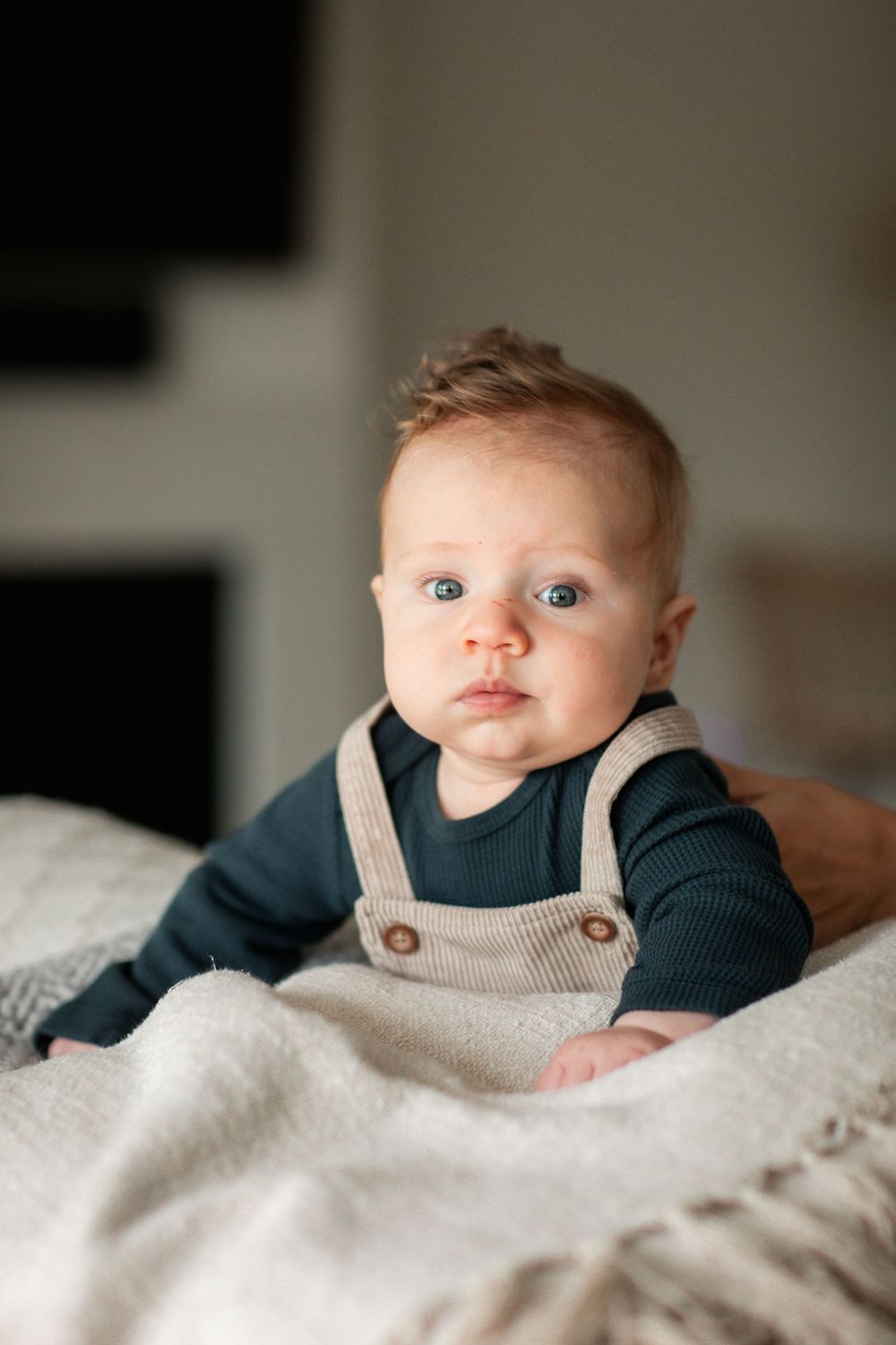 Adorable baby with blue eyes wearing a dark teal onesie and beige overalls, lying on a white blanket.