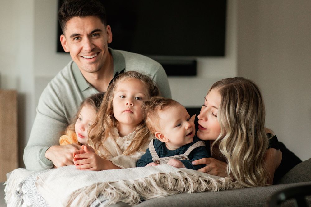 Happy family of five posing together on a couch, parents smiling with three young children in a bright modern home.