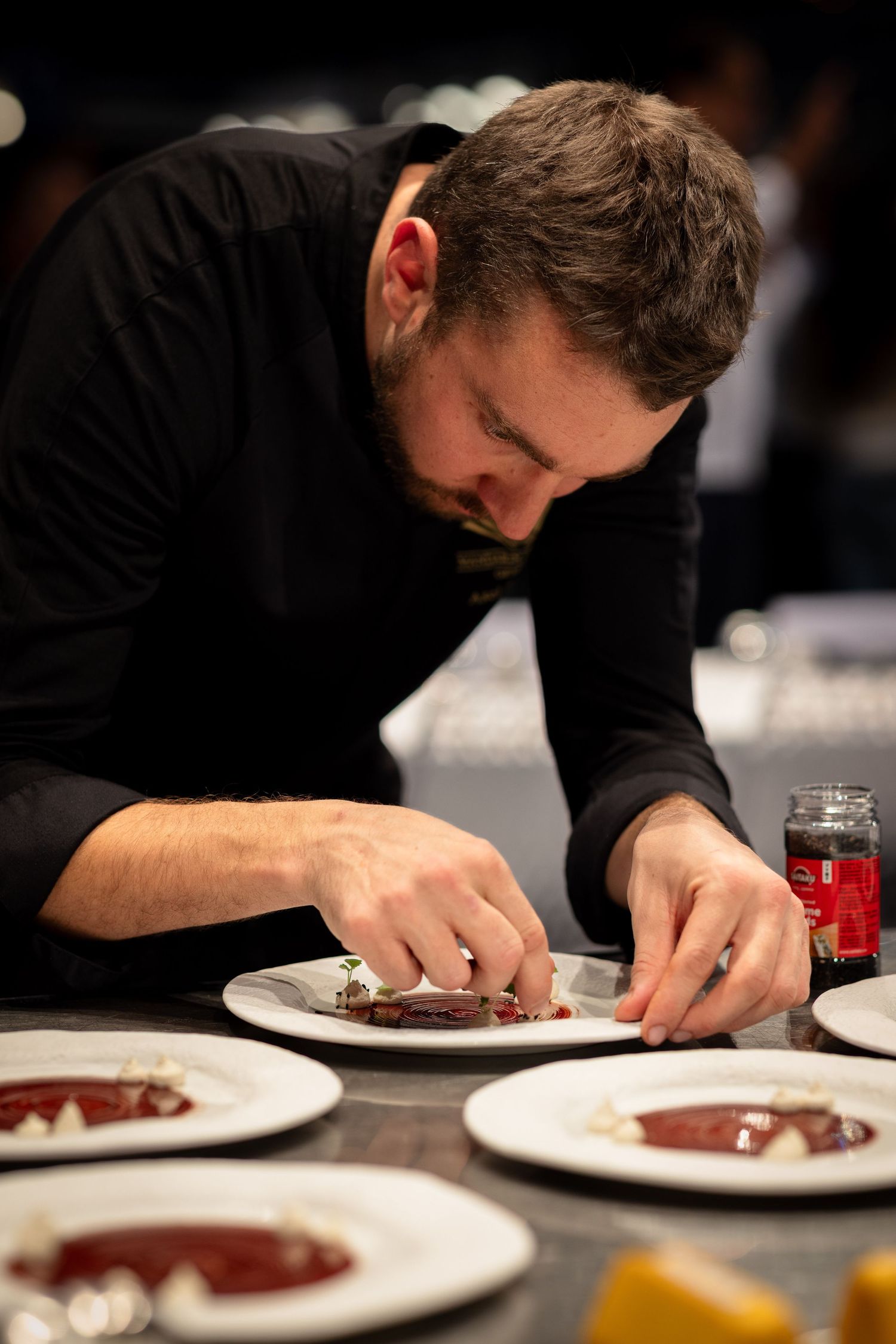 Chef in black attire carefully plating and preparing food on white dishes at a restaurant counter.