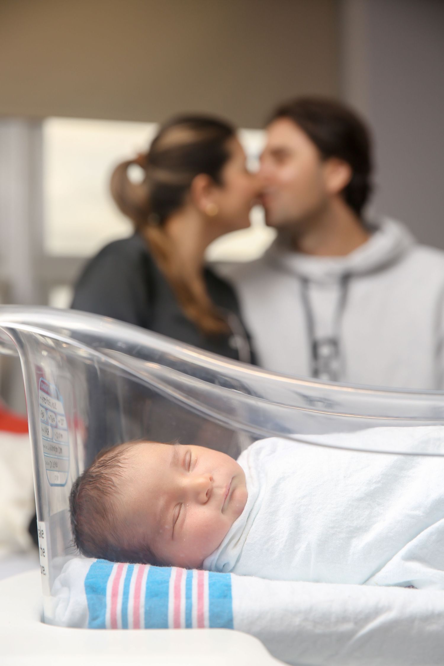 A red-haired woman with glasses tenderly holds a sleeping newborn baby in a hospital room.