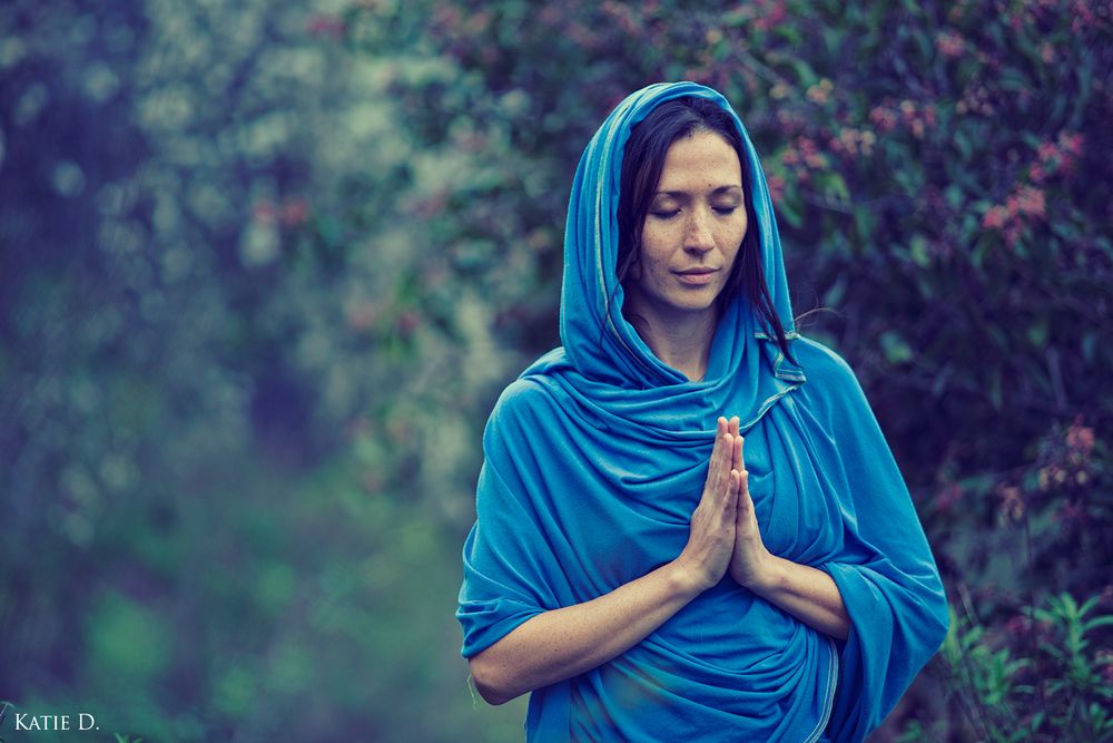 Serene woman in blue hooded shawl meditating with hands in namaste gesture outdoors.