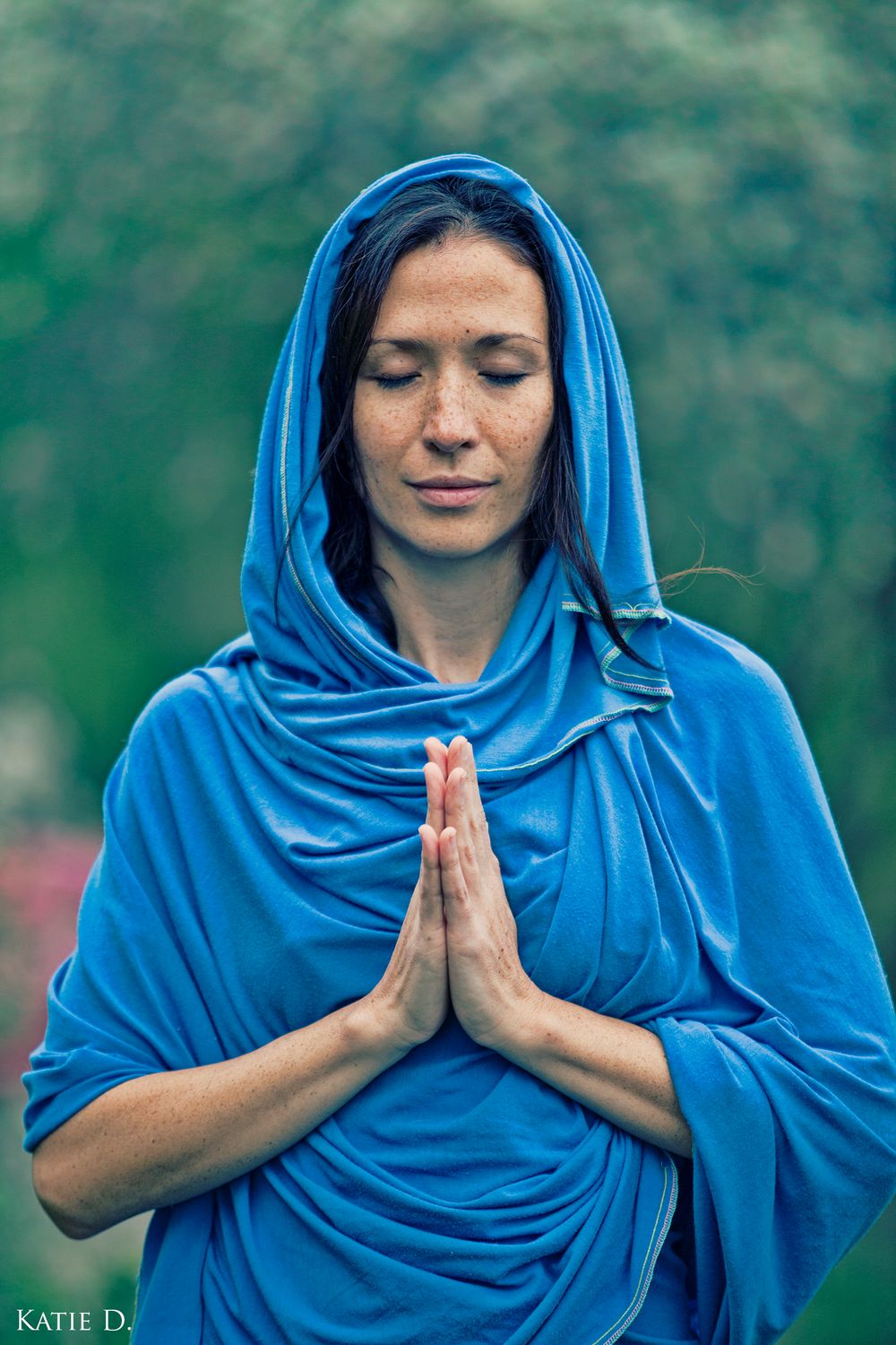 Close-up of woman in blue hood eyes closed hands in prayer pose in nature.