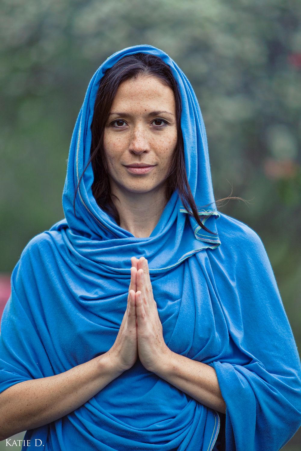 Woman with freckles in blue hooded shawl holding prayer hands looking at camera outdoors.
