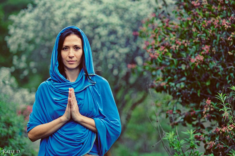 Woman in teal hooded wrap with hands in namaste gesture standing in green garden.