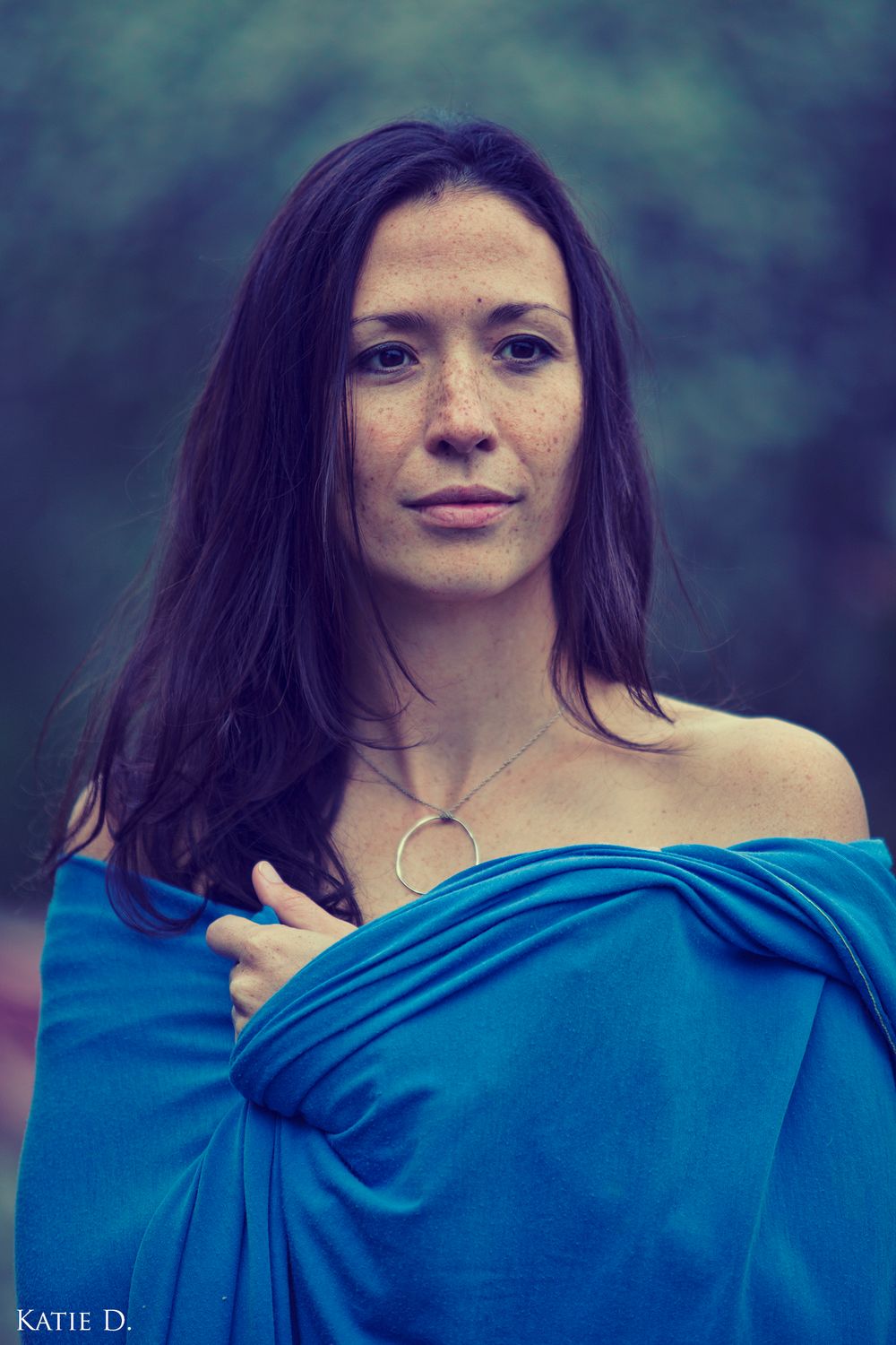Portrait of brunette woman with freckles in off-shoulder blue wrap wearing circle necklace.