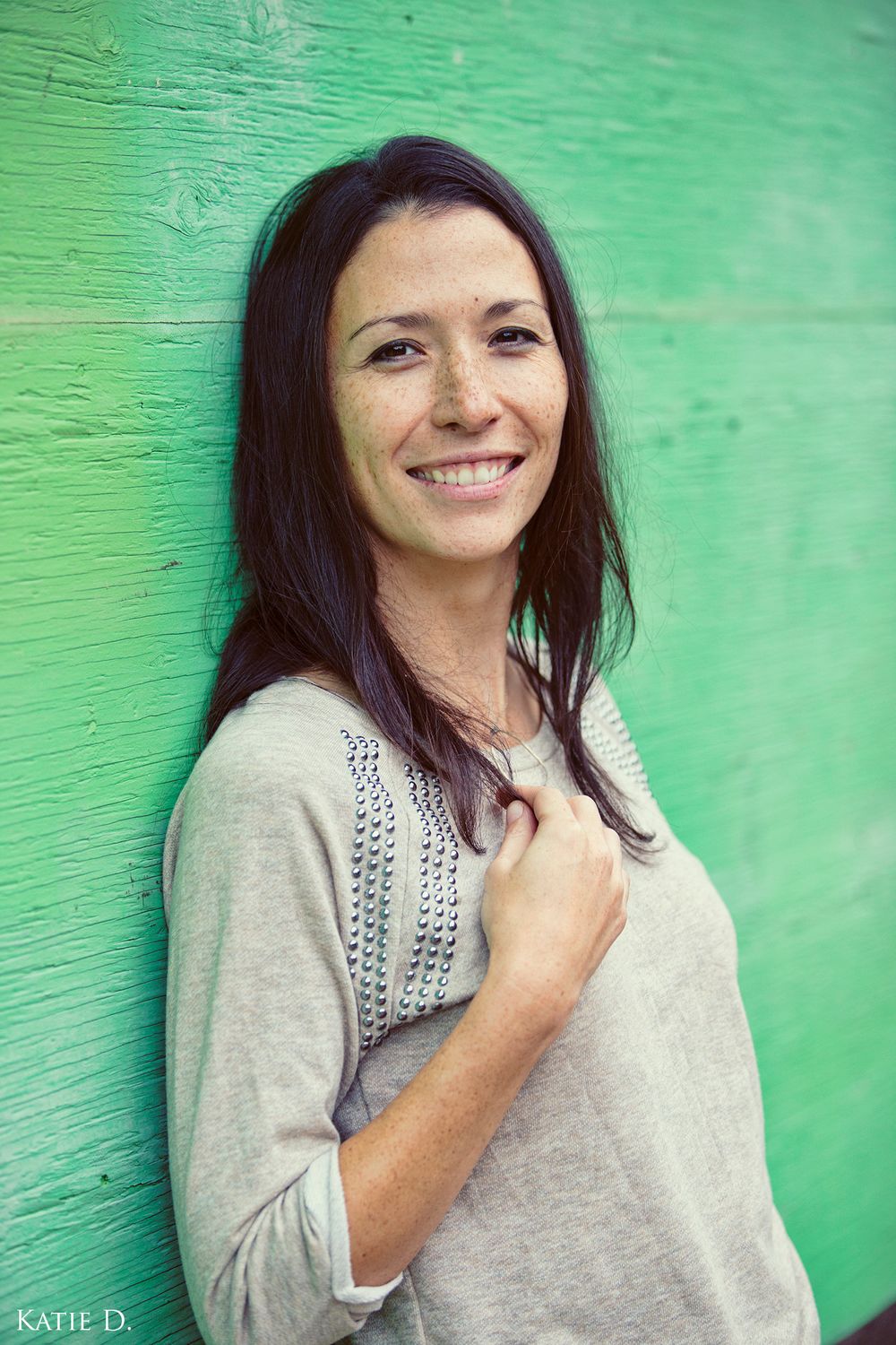 Smiling dark-haired woman in gray knit sweater posing against bright green wall.