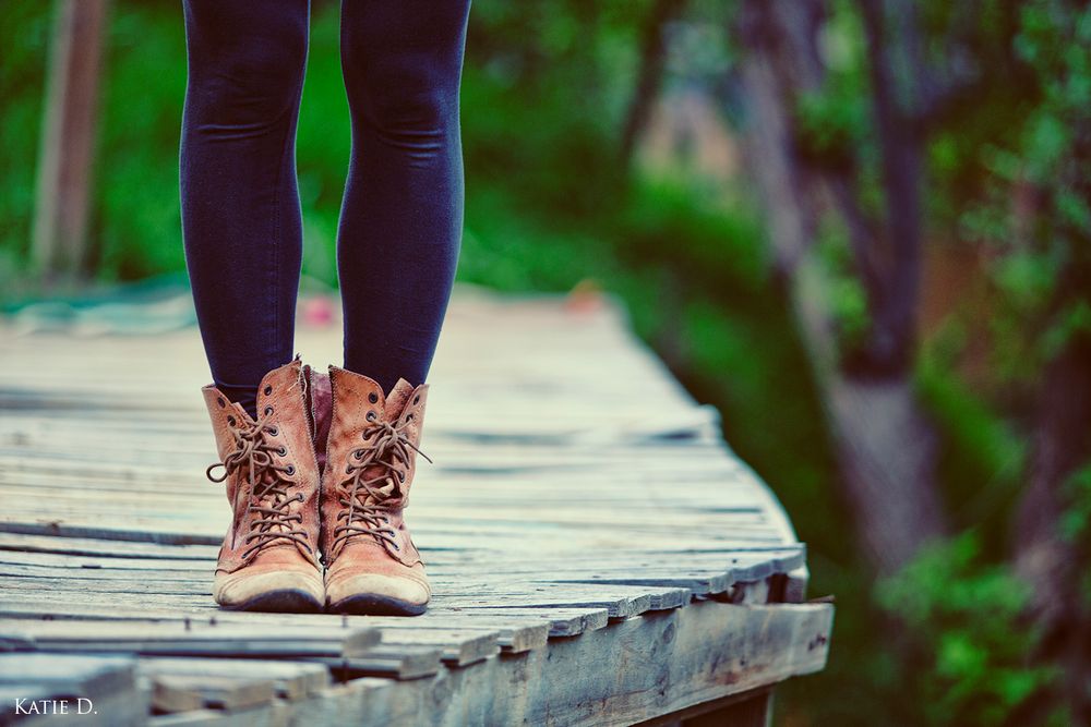 Brown lace-up ankle boots and black leggings on weathered wooden bridge in green forest setting.