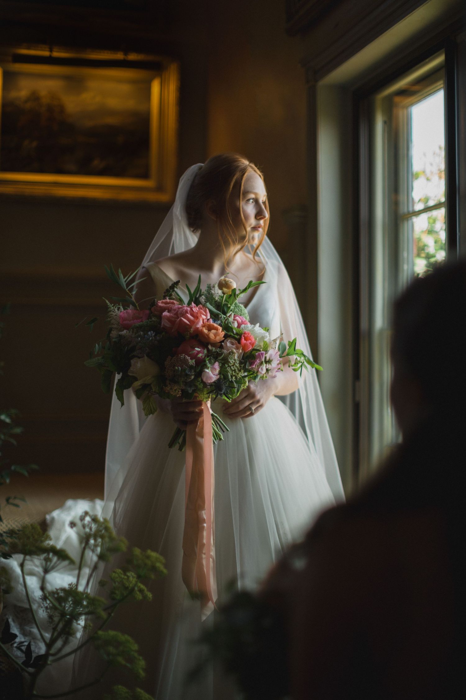 Bride in white gown and veil holds colorful bouquet with pink ribbon, standing by window in elegant room.