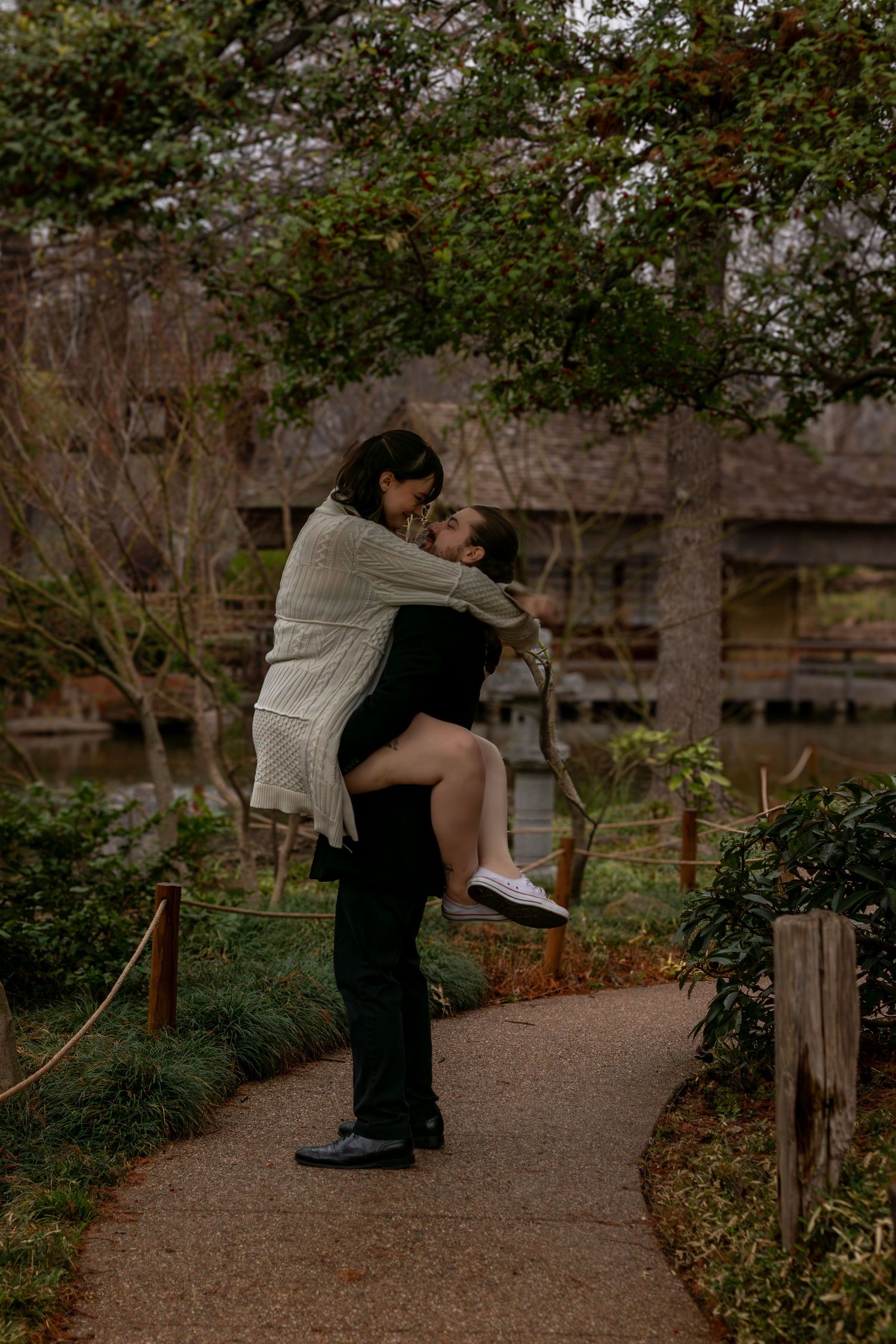 A couple shares a romantic kiss as the man lifts the woman on a garden path surrounded by lush greenery.