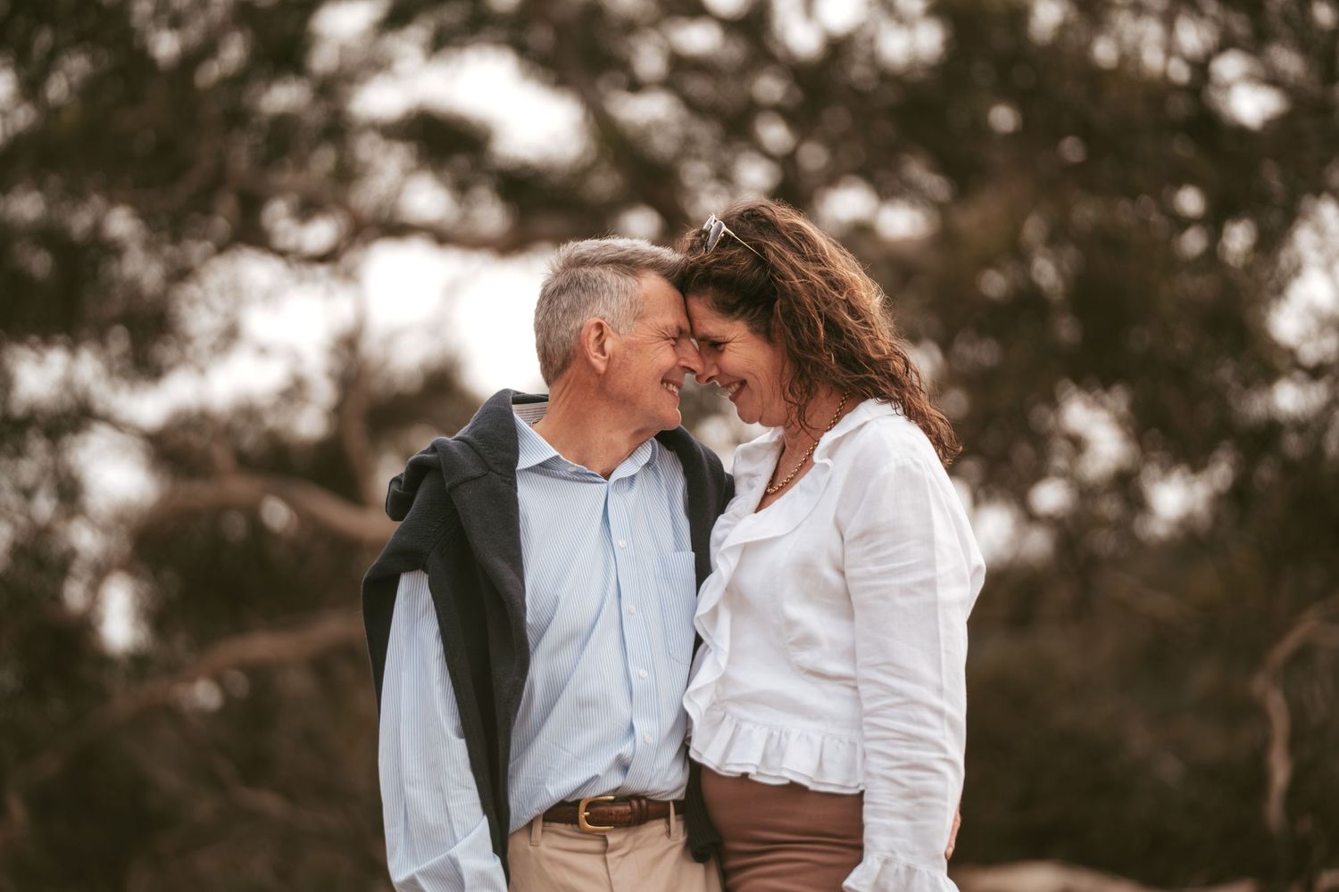 A middle-aged couple shares a romantic moment outdoors, smiling at each other with trees in the background.