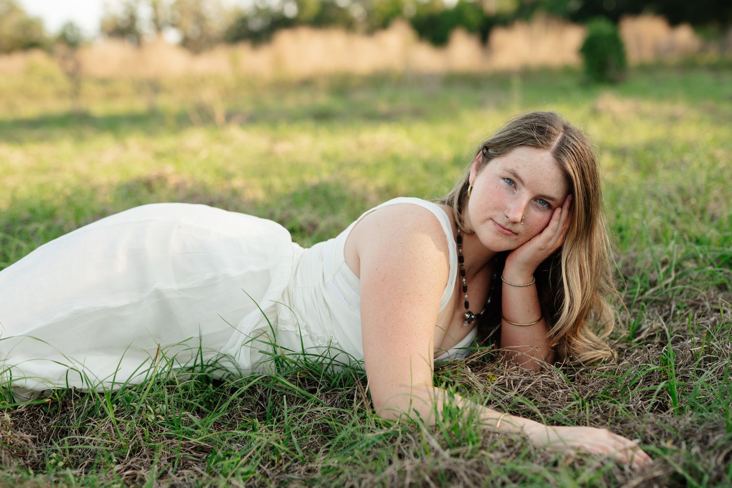 A young woman in a white dress lies on the grass with her head resting on her hand, smiling softly during an outdoor portrait session.