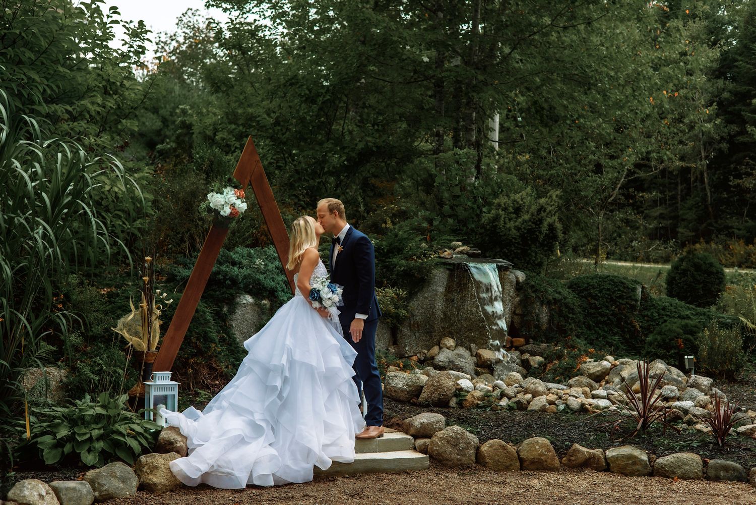 Bride and groom kissing in a lush garden wedding ceremony with a rustic wooden arch and waterfall backdrop.