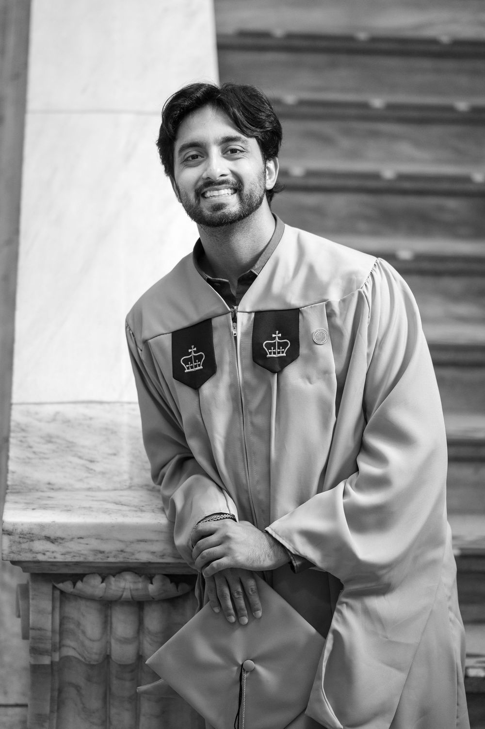 Young man in graduation gown smiling while leaning on marble railing in black and white photo.