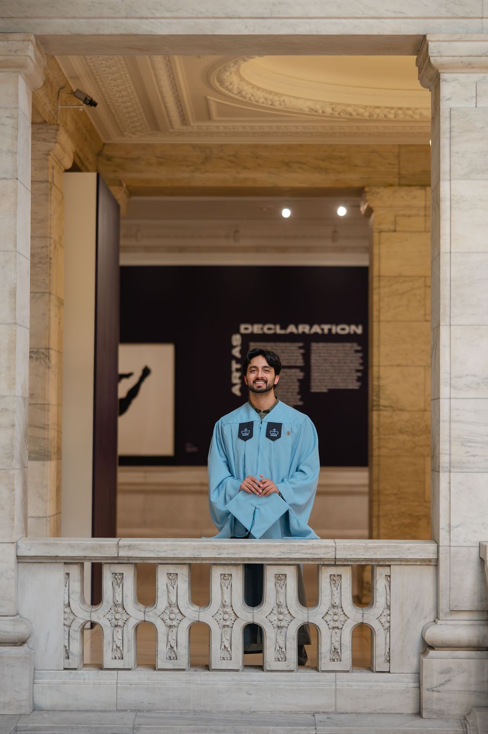 Man in light blue traditional attire smiling on a balcony inside a grand museum near a Declaration exhibit.